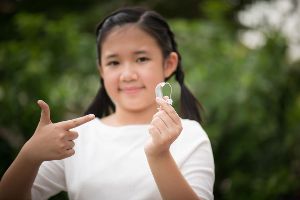 A young girl holds up her hearing aid and gives a thumbs up.